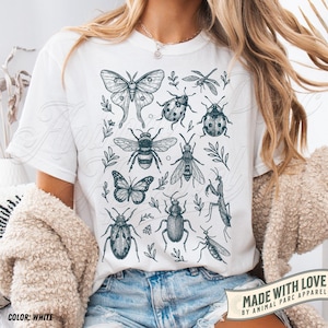 May include: White t-shirt with a detailed illustration of insects, such as a moth, bee, butterfly, and beetles, with leafy accents. The shirt has the text "MADE WITH LOVE BY ANIMAL PARC APPAREL" and "COLOR: WHITE".