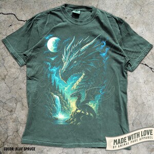 May include: Blue spruce t-shirt featuring a detailed dragon illustration with a full moon, waterfall, and forest scene. The dragon is rendered in shades of blue and white, with a detailed design. The text "MADE WITH LOVE BY ANIMAL PARC APPAREL" is printed on the bottom right.