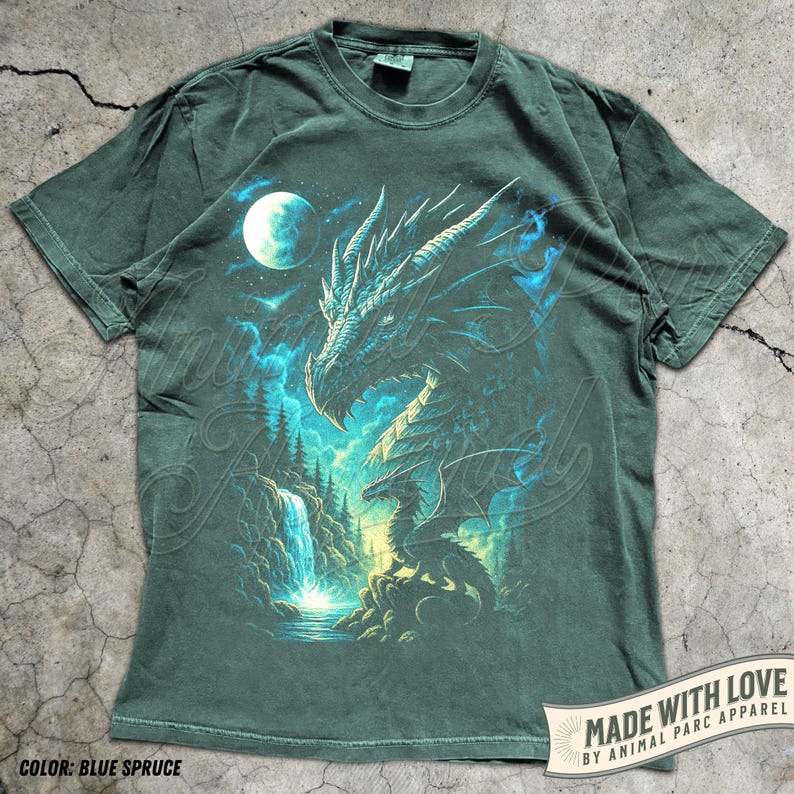 May include: Blue spruce t-shirt featuring a detailed dragon illustration with a full moon, waterfall, and forest scene. The dragon is rendered in shades of blue and white, with a detailed design. The text "MADE WITH LOVE BY ANIMAL PARC APPAREL" is printed on the bottom right.
