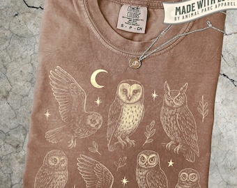 May include: Espresso-colored t-shirt featuring a detailed illustration of several owls, a crescent moon, and stars. The shirt has a crew neck and a tag that reads "COMFORT COLORS". A silver necklace with a pendant is draped across the shirt. The text "MADE WITH LOVE" is also visible.