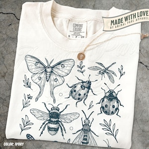 May include: Cream-coloured t-shirt with a dark teal insect illustration, including a moth, dragonfly, bee, and ladybug, plus sprigs of leaves. The shirt has a crew neck and short sleeves. The text "MADE WITH LOVE BY ANIMAL PARC APPAREL" is visible.