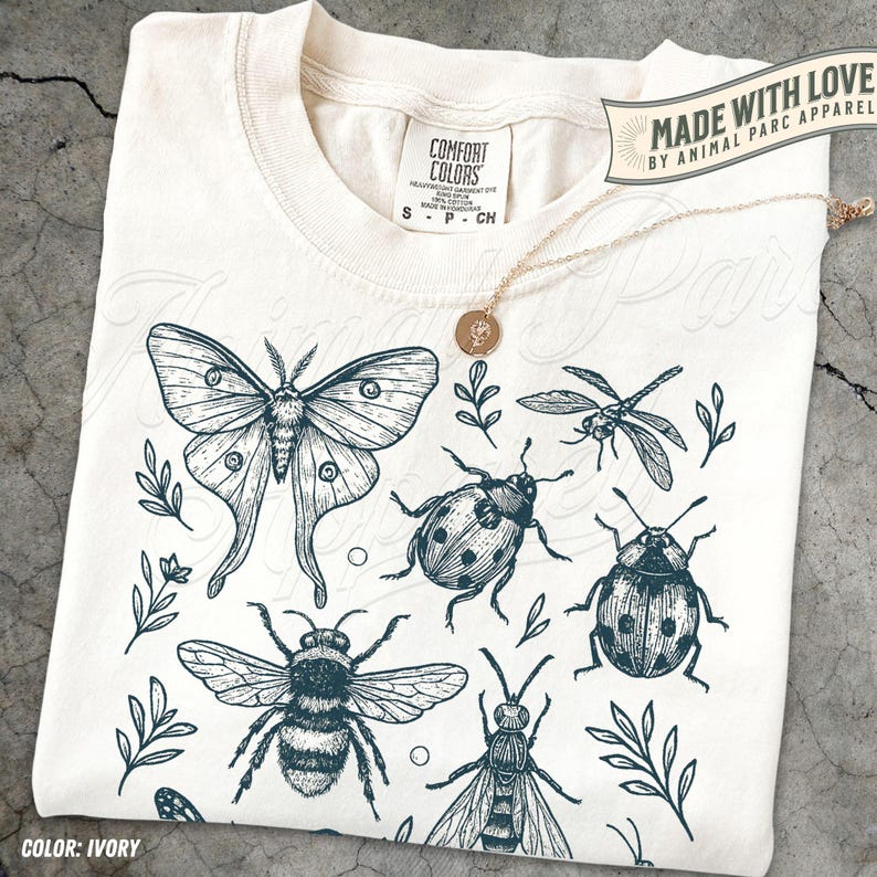May include: Cream-coloured t-shirt with a dark teal insect illustration, including a moth, dragonfly, bee, and ladybug, plus sprigs of leaves. The shirt has a crew neck and short sleeves. The text "MADE WITH LOVE BY ANIMAL PARC APPAREL" is visible.