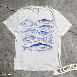 May include: White t-shirt featuring a blue illustration of various fish species. The design includes detailed drawings of different fish, with their names written in cursive. The shirt has a crew neck and short sleeves. The text "MADE WITH LOVE BY ANIMAL PARC APPAREL" is printed on the bottom right.
