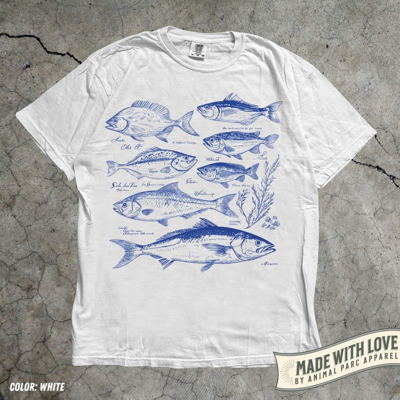 May include: White t-shirt featuring a blue illustration of various fish species. The design includes detailed drawings of different fish, with their names written in cursive. The shirt has a crew neck and short sleeves. The text "MADE WITH LOVE BY ANIMAL PARC APPAREL" is printed on the bottom right.