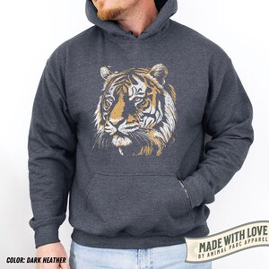 May include: Dark heather gray hoodie featuring a tiger face graphic in shades of gold and white. The hoodie has a kangaroo pocket and a drawstring hood. The text "MADE WITH LOVE BY ANIMAL PARC APPAREL" is printed on a white banner.