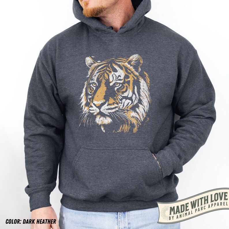 May include: Dark heather gray hoodie featuring a tiger face graphic in shades of gold and white. The hoodie has a kangaroo pocket and a drawstring hood. The text "MADE WITH LOVE BY ANIMAL PARC APPAREL" is printed on a white banner.