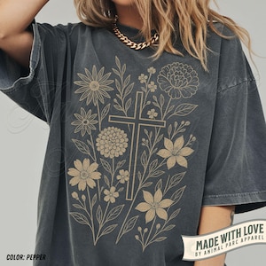 May include: Pepper-coloured t-shirt with a floral design and a cross at the centre. The design includes various flowers and leaves in a light brown colour. The shirt has short sleeves and a relaxed fit. The text "MADE WITH LOVE" is printed on the bottom right.