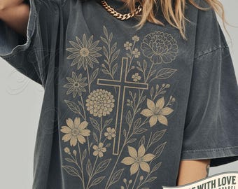 Könnte beinhalten: Pfefferfarbenes T-Shirt mit floralem Design und einem Kreuz in der Mitte. Das Design umfasst verschiedene Blumen und Blätter in Hellbraun. Das Shirt hat kurze Ärmel und eine lockere Passform. Der Text "MADE WITH LOVE" ist unten rechts aufgedruckt.