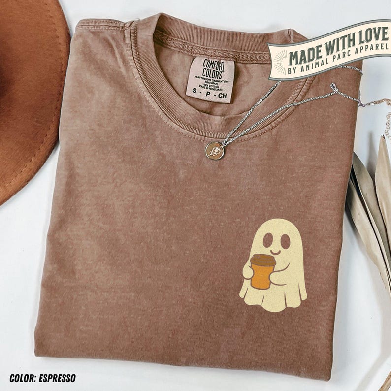 May include: Espresso-colored Comfort Colors t-shirt with a ghost graphic holding a coffee cup. The shirt has a small tag and a silver necklace. The text "MADE WITH LOVE BY ANIMAL PARC APPAREL" is visible.