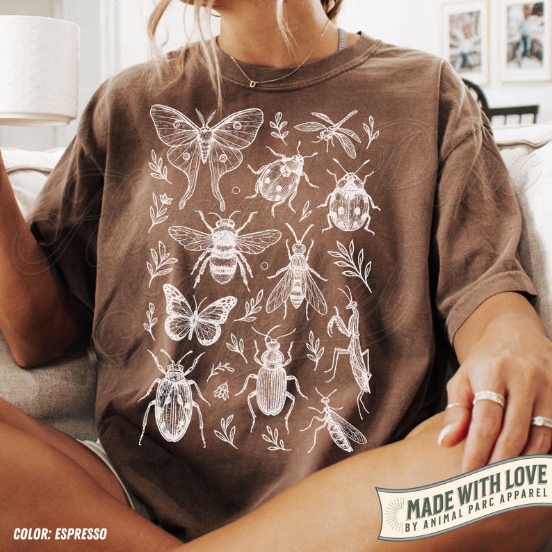 May include: Espresso-coloured t-shirt with a white insect and botanical illustration. The design includes a moth, butterfly, bee, ladybug, and other insects, along with sprigs of leaves. The shirt has a relaxed fit and is made with love by Animal Parc Apparel.
