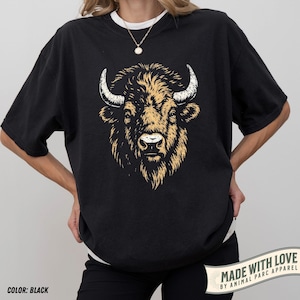 May include: Black t-shirt featuring a detailed illustration of a bison's head. The bison has tan fur and white horns. The text "MADE WITH LOVE BY ANIMAL PARC APPAREL" is printed on a banner on the lower right. The shirt is black and the color is noted as "COLOR: BLACK".
