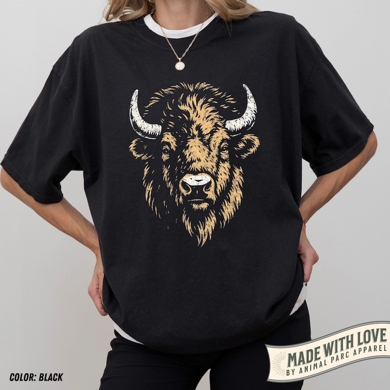 May include: Black t-shirt featuring a detailed illustration of a bison's head. The bison has tan fur and white horns. The text "MADE WITH LOVE BY ANIMAL PARC APPAREL" is printed on a banner on the lower right. The shirt is black and the color is noted as "COLOR: BLACK".