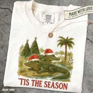 May include: Ivory t-shirt with a Christmas-themed design. The graphic shows two alligators in Santa hats, Christmas trees, and a palm tree. The text "'TIS THE SEASON" is printed in red. The shirt also has the text "MADE WITH LOVE BY ANIMAL PARC APPAREL".
