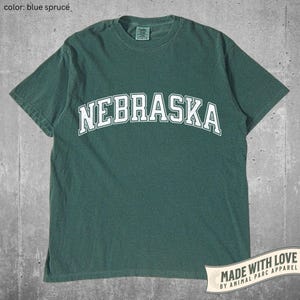 Könnte beinhalten: Blau-Fichte-T-Shirt mit dem Wort "NEBRASKA" in weißen, gewölbten Buchstaben. Das Shirt hat einen Rundhalsausschnitt und kurze Ärmel. Ein Banner unten rechts lautet "MADE WITH LOVE BY ANIMAL PARC APPAREL."