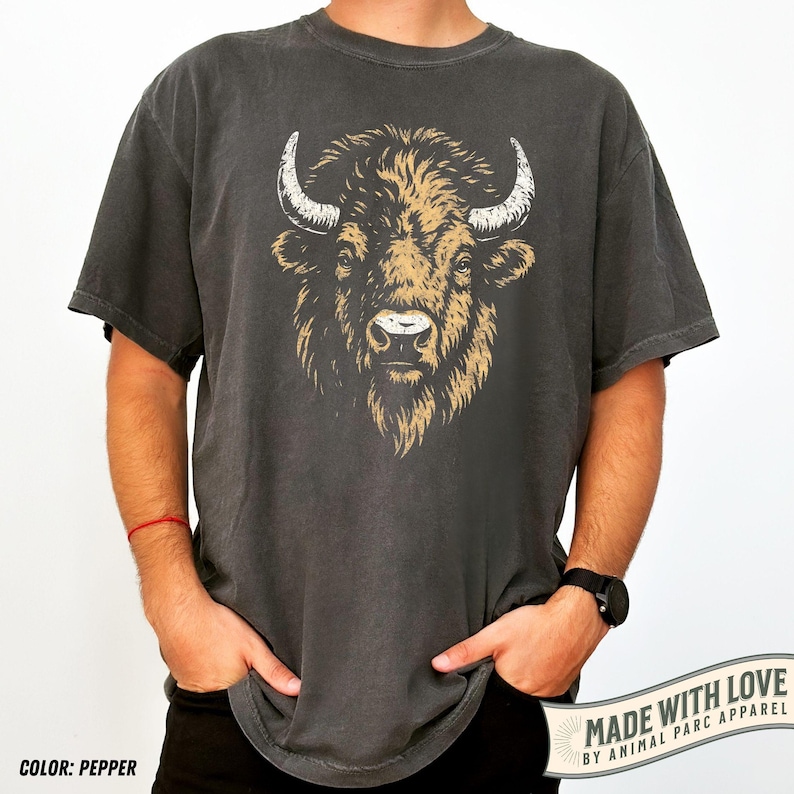 May include: Pepper-colored t-shirt featuring a detailed illustration of a bison's head with large horns. The bison is depicted in shades of brown and tan against a dark background. The text "MADE WITH LOVE BY ANIMAL PARC APPAREL" is printed on the bottom right.