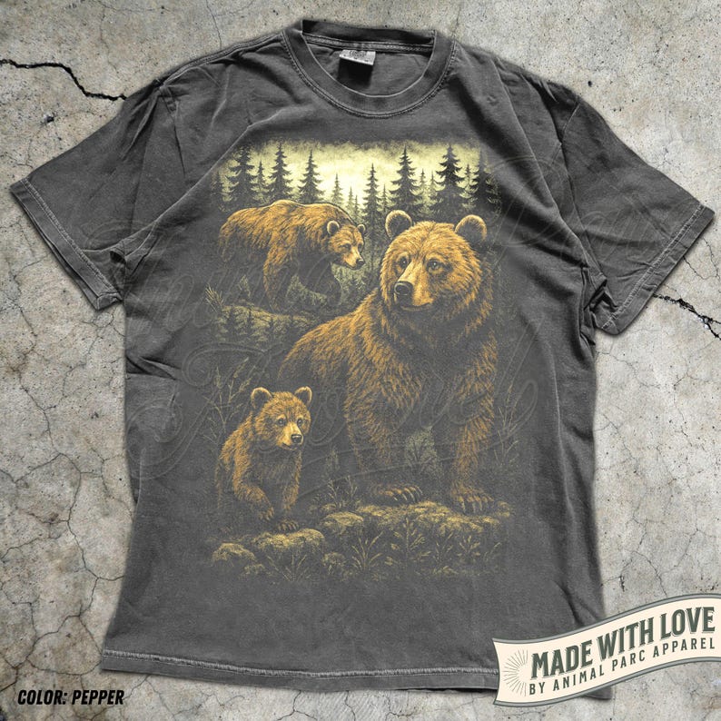 May include: Pepper-colored t-shirt featuring a detailed illustration of three brown bears in a forest setting. The design includes a mother bear and two cubs, with a backdrop of pine trees. The shirt has a classic crew neck and short sleeves.