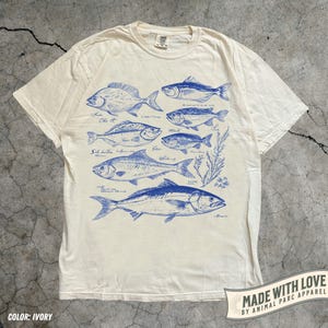 May include: Ivory-colored t-shirt featuring blue illustrations of various fish species, each labeled with their names. The design includes detailed drawings of the fish and some aquatic plants. The shirt has a classic crew neck and short sleeves.