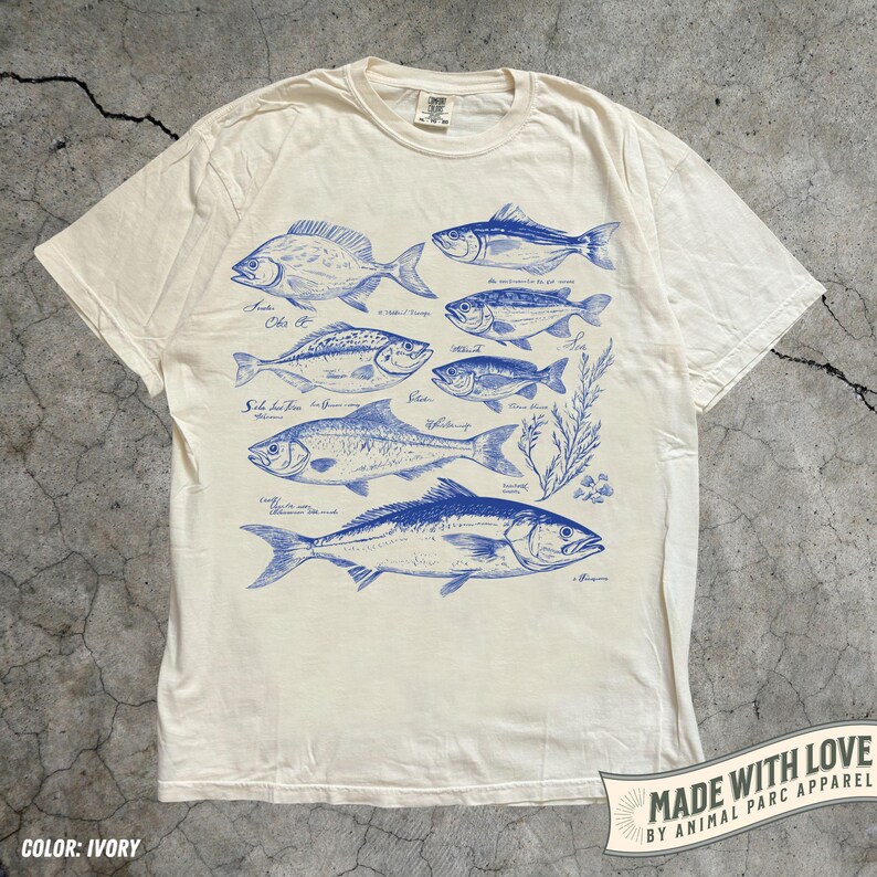 May include: Ivory-colored t-shirt featuring blue illustrations of various fish species, each labeled with their names. The design includes detailed drawings of the fish and some aquatic plants. The shirt has a classic crew neck and short sleeves.