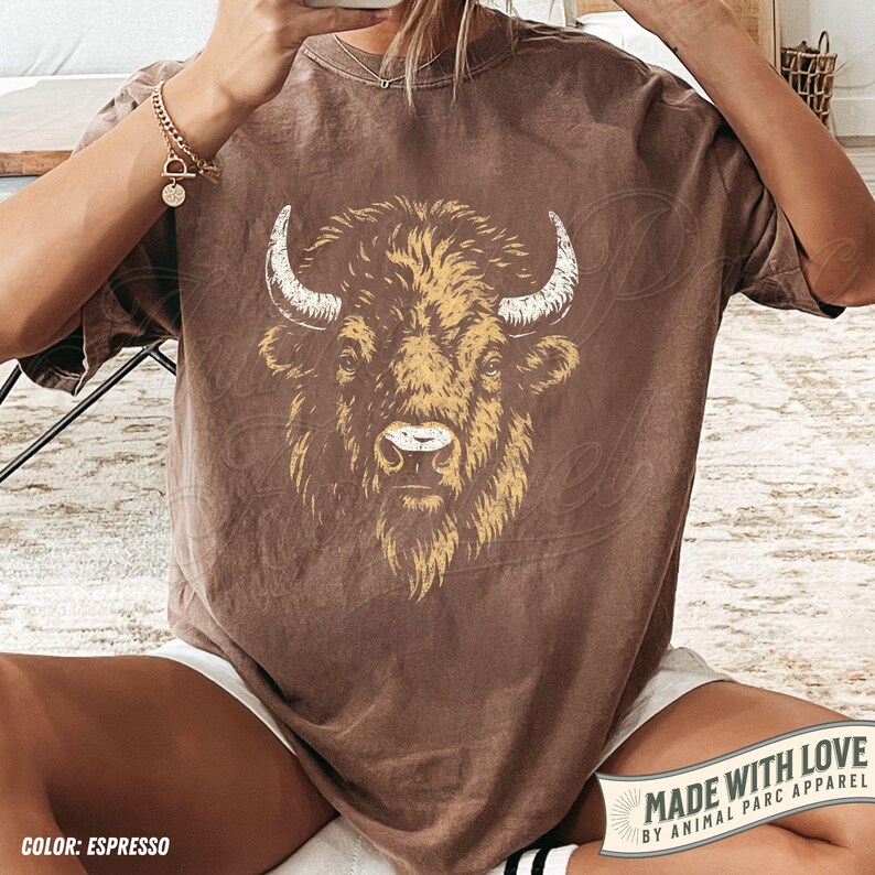 May include: Espresso-colored t-shirt featuring a detailed illustration of a bison's head. The bison is depicted with white horns and a yellow-toned face, set against a brown background. The shirt's color is labeled as "Espresso".