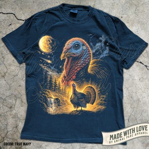 Puede incluir: Una camiseta de manga corta azul marino con un estampado gr&aacute;fico de una cabeza de pavo grande y un pavo m&aacute;s peque&ntilde;o en un paisaje forestal con una cascada, una luna amarilla brillante y un cielo estrellado. El texto dice Made with love by Animal Parc Apparel.