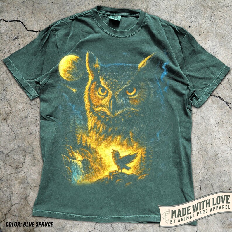 May include: Blue spruce t-shirt with a detailed illustration of an owl with yellow eyes, a full moon, and a singing bird. The design includes a waterfall and forest scene. The shirt has the text "MADE WITH LOVE BY ANIMAL PARC APPAREL".