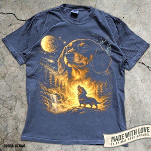 May include: Denim blue t-shirt featuring a detailed illustration of a dachshund dog under a full moon, with a waterfall and forest scene. The shirt includes the text "MADE WITH LOVE BY ANIMAL PARC APPAREL" and "COLOR: DENIM".