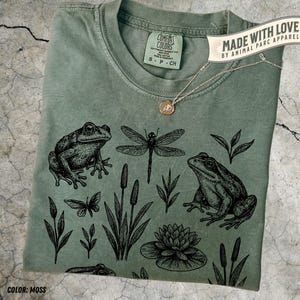 May include: A moss-colored t-shirt with a nature-themed design featuring frogs, a dragonfly, and aquatic plants. The shirt includes the text "COMFORT COLORS" and "MADE WITH LOVE BY ANIMAL PARC APPAREL". A gold necklace with a pendant is also visible.
