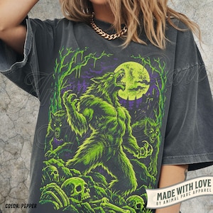 Puede incluir: Una camiseta gris carbón con un estampado gráfico de un hombre lobo grande aullando a una luna amarilla brillante, rodeado de dos lobos más pequeños, árboles desnudos y cráneos humanos esparcidos por el suelo. Corte holgado y cuadrado.