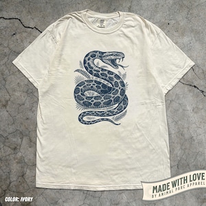 Könnte beinhalten: Elfenbeinfarbenes T-Shirt mit einer dunkelblauen Illustration einer zusammengerollten Schlange mit herausgestreckter Zunge, umgeben von Blättern. Das Shirt hat einen Rundhalsausschnitt und kurze Ärmel. Der Text "MADE WITH LOVE BY ANIMAL PARC APPAREL" ist auf einem Banner gedruckt.