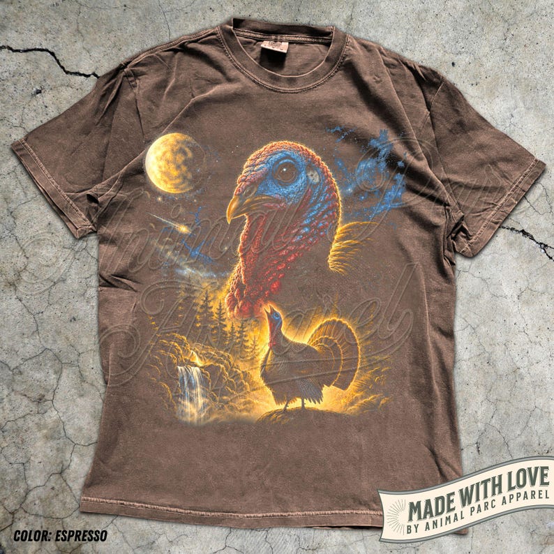 Puede incluir: Una camiseta de manga corta color marr&oacute;n espresso con un estampado gr&aacute;fico de la cabeza de un pavo y un pavo de cuerpo entero junto a una cascada y un bosque. El dise&ntilde;o incluye una luna amarilla, una estrella fugaz y nubes azules.