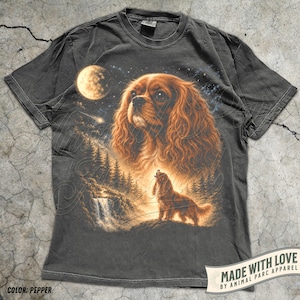 Op de afbeelding: Pepperkleurig t-shirt met een gedetailleerde illustratie van een Cavalier King Charles Spaniel onder een sterrenhemel met volle maan. Het ontwerp bevat een waterval en dennenbomen, met een tweede huilende hond. De tekst "MADE WITH LOVE" is zichtbaar.