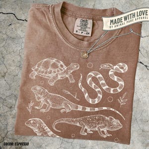 Peut inclure: T-shirt couleur expresso avec des illustrations blanches de reptiles et d'amphibiens, dont une tortue, un serpent, un iguane et un caméléon. Le t-shirt a un col rond et une petite étiquette indiquant "Comfort Colors". Le texte "MADE WITH LOVE BY ANIMAL PARC APPAREL" est également visible.