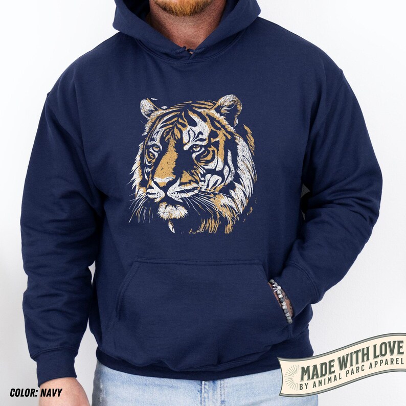 May include: Navy blue hoodie featuring a detailed tiger face graphic in shades of gold and white. The hoodie has a kangaroo pocket and a drawstring hood. The text "MADE WITH LOVE BY ANIMAL PARC APPAREL" is printed on a banner.