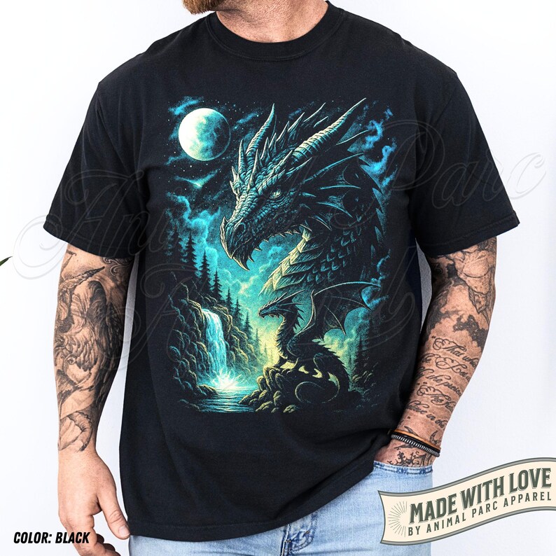 May include: Black t-shirt featuring a detailed dragon graphic in shades of teal and blue, set against a night sky with a full moon and waterfall. The text "MADE WITH LOVE BY ANIMAL PARC APPAREL" is printed on the bottom right.