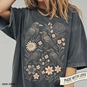 Pode incluir: T-shirt cor pimenta com uma ilustração detalhada de quatro corvos entre elementos florais. O design inclui várias flores e folhas em uma cor complementar. A camisa tem um ajuste folgado e mangas curtas. O texto "MADE WITH LOVE BY ANIMAL PARC APPAREL" é visível.