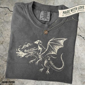 May include: A gray Comfort Colors t-shirt with a cream-colored graphic of a dog riding a dragon, with the text "MADE WITH LOVE BY ANIMAL PARC APPAREL". A silver necklace with a pendant is draped on the shirt. The shirt color is Pepper.