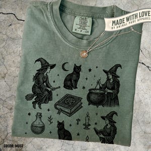 Può includere: T-shirt verde muschio con grafica a tema streghe. Il design include streghe, gatti neri, un calderone, un libro degli incantesimi e una bottiglia di pozione. La maglietta ha il testo "COMFORT COLORS" e "MADE WITH LOVE BY ANIMAL PARC APPAREL".