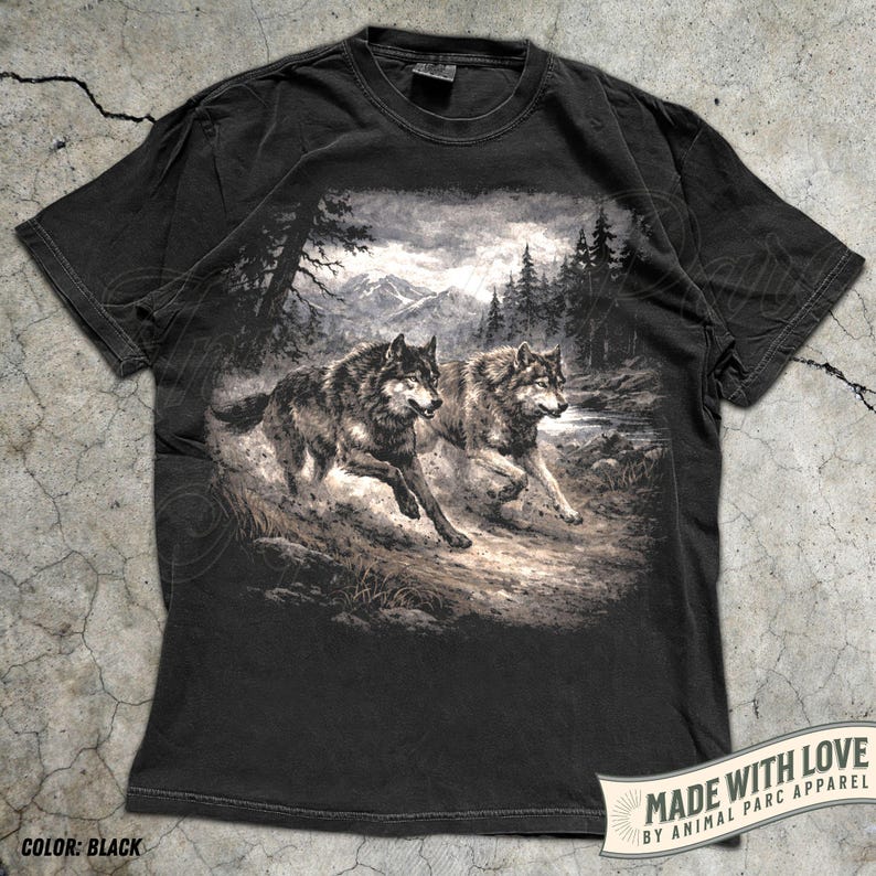 K&ouml;nnte beinhalten: Schwarzes T-Shirt mit einer detaillierten Grafik von zwei W&ouml;lfen, die durch eine Waldlandschaft mit Bergen im Hintergrund laufen. Das Shirt hat einen Used-Look und den Text "MADE WITH LOVE BY ANIMAL PARC APPAREL".
