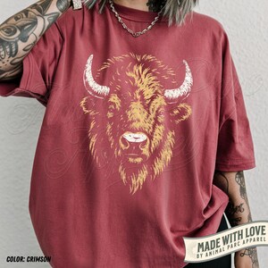 May include: Crimson-colored t-shirt featuring a detailed illustration of a bison's head with white horns and a golden-yellow mane. The text "MADE WITH LOVE BY ANIMAL PARC APPAREL" is printed on a banner. The shirt's color is labeled "COLOR: CRIMSON".