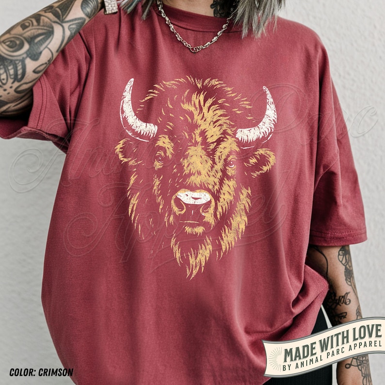 May include: Crimson-colored t-shirt featuring a detailed illustration of a bison's head with white horns and a golden-yellow mane. The text "MADE WITH LOVE BY ANIMAL PARC APPAREL" is printed on a banner. The shirt's color is labeled "COLOR: CRIMSON".