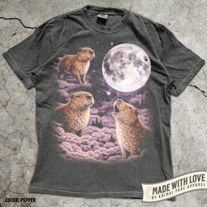 May include: Pepper-colored t-shirt featuring a graphic of capybaras under a full moon. The design includes three capybaras, clouds, and a starry night sky. The shirt has a crew neck and short sleeves. The text "MADE WITH LOVE BY ANIMAL PARC APPAREL" is also visible.