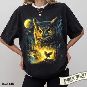 May include: Black t-shirt with a detailed illustration of an owl with yellow eyes, set against a moonlit forest scene. The design includes a small bird and the text "MADE WITH LOVE BY ANIMAL PARC APPAREL".