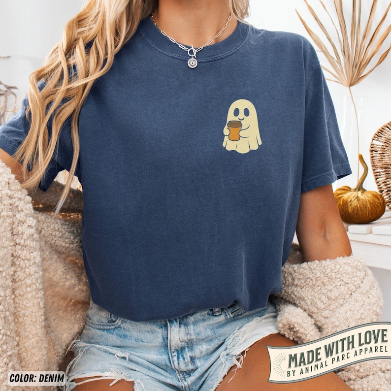 May include: Denim blue t-shirt featuring a small, smiling ghost holding a coffee cup. The shirt has a crew neck and short sleeves. The text "MADE WITH LOVE BY ANIMAL PARC APPAREL" is printed on a banner at the bottom right.