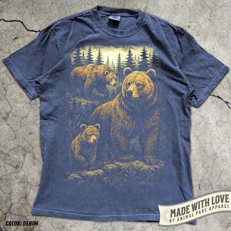 May include: Denim blue t-shirt featuring a detailed illustration of a bear family in a forest setting. The design includes a large bear with two cubs, set against a backdrop of trees. The shirt has the text "MADE WITH LOVE BY ANIMAL PARC APPAREL".