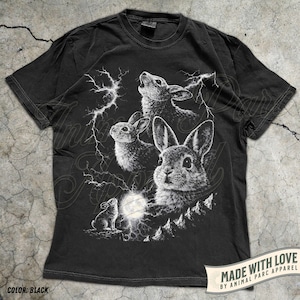 Pode incluir: T-shirt preto com uma ilustra&ccedil;&atilde;o branca de coelhos, raios e lua cheia. O design cria um contraste marcante no tecido escuro. A camisa inclui o texto "MADE WITH LOVE BY ANIMAL PARC APPAREL" e "COLOR: BLACK".