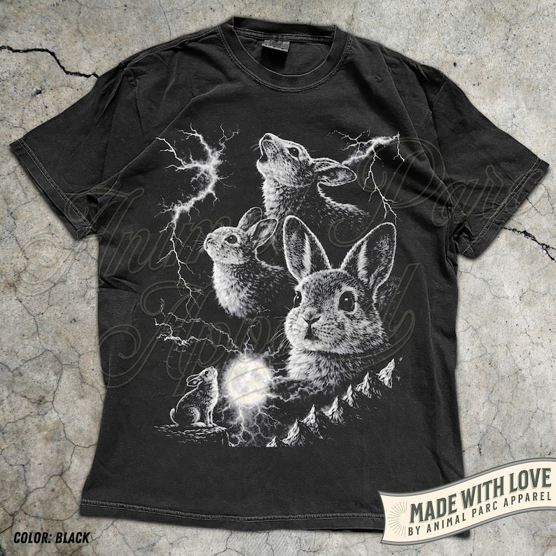 Pode incluir: T-shirt preto com uma ilustra&ccedil;&atilde;o branca de coelhos, raios e lua cheia. O design cria um contraste marcante no tecido escuro. A camisa inclui o texto "MADE WITH LOVE BY ANIMAL PARC APPAREL" e "COLOR: BLACK".
