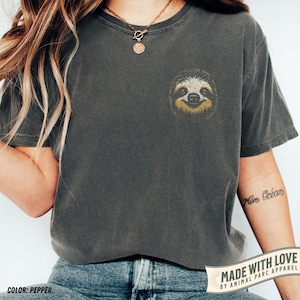 May include: Pepper-coloured t-shirt featuring an embroidered sloth face on the chest. The sloth has a yellow and brown face and a smiling expression. The shirt is a relaxed fit with short sleeves. The text "MADE WITH LOVE BY ANIMAL PARC APPAREL" is printed on a banner.