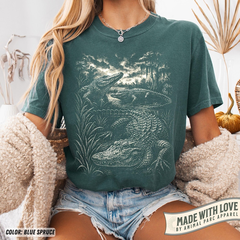 May include: Blue spruce t-shirt featuring a detailed illustration of alligators in a swamp setting. The graphic is in a vintage style, with the text "MADE WITH LOVE BY ANIMAL PARC APPAREL" at the bottom. The shirt is a comfortable, short-sleeved style.