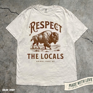 May include: Ivory-colored t-shirt with a brown graphic of a bison and the text "RESPECT THE LOCALS" above and below the image. The shirt also includes the text "ANIMAL PARC AP." and "MADE WITH LOVE BY ANIMAL PARC APPAREL".