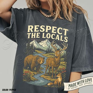 May include: Pepper-colored t-shirt with a nature-themed graphic. The design features a bear, deer, and fox near a river, with mountains in the background. The text "RESPECT THE LOCALS" is printed above the illustration. The shirt also has the text "MADE WITH LOVE BY ANIMAL PARC APPAREL".
